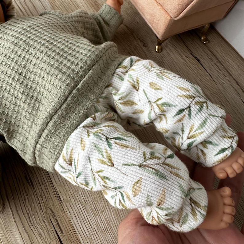 Pu&ograve; includere: Una bambola indossa una maglia a maniche lunghe in tessuto waffle color verde salvia e pantaloni a coste bianchi con un motivo di piccole foglie verdi e marroni. L'abbigliamento presenta polsini e orli, con la trama del tessuto ben visibile.