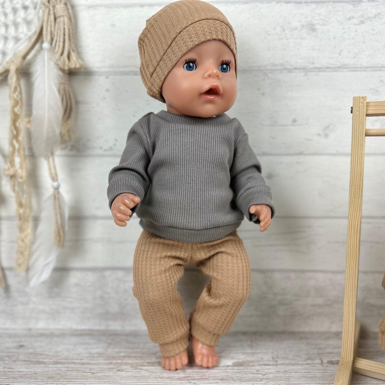 May include: A doll wearing a tan waffle-knit beanie, a long-sleeved grey ribbed shirt, and matching tan waffle-knit trousers. The doll has blue eyes and a neutral expression, with the clothing featuring a consistent textured fabric design.