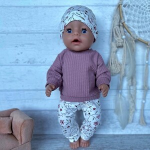 May include: A doll wearing a matching set consisting of a white beanie with a green leaf pattern, a long-sleeved mauve ribbed shirt, and white leggings featuring a delicate floral pattern with small red and green flowers and thin stems.