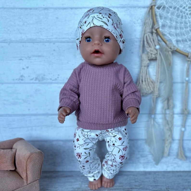 May include: A doll wearing a matching set consisting of a white beanie with a green leaf pattern, a long-sleeved mauve ribbed shirt, and white leggings featuring a delicate floral pattern with small red and green flowers and thin stems.