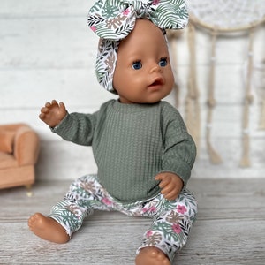 May include: A doll wearing a green waffle knit long-sleeve shirt and white pants with a pink and green floral print. The doll is wearing a headband with a pink and green floral print.