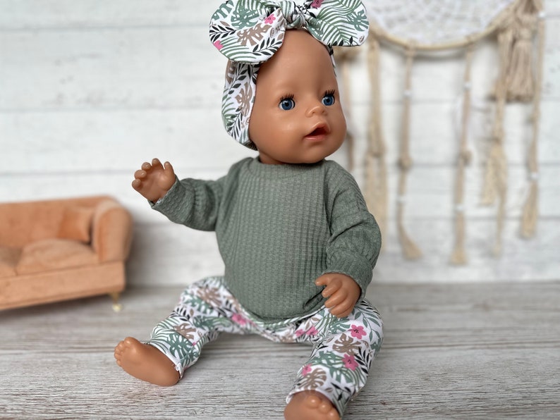 May include: A doll wearing a green waffle knit long-sleeve shirt and white pants with a pink and green floral print. The doll is wearing a headband with a pink and green floral print.