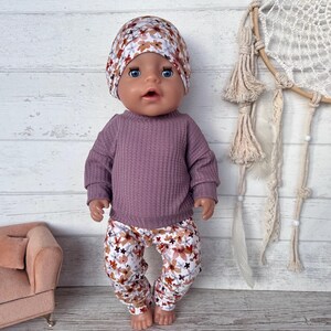 May include: A doll dressed in a mauve waffle-knit top, floral print trousers, and a matching beanie. The trousers and beanie have a pattern of brown, white, and blue flowers. A small pink sofa is in the background.