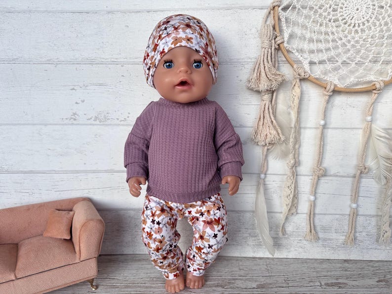 May include: A doll dressed in a mauve waffle-knit top, floral print trousers, and a matching beanie. The trousers and beanie have a pattern of brown, white, and blue flowers. A small pink sofa is in the background.