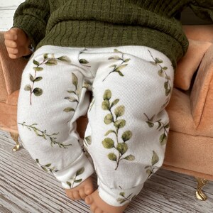 May include: White baby leggings with a green leafy print. The leggings are on a small peach-coloured sofa.