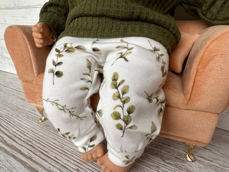 May include: White baby leggings with a green leafy print. The leggings are on a small peach-coloured sofa.