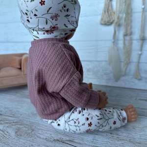 May include: A doll wearing a dusty rose ribbed long-sleeve shirt and white leggings featuring a delicate floral pattern of small red flowers and thin green vines. The doll also wears a matching white beanie with the same floral print design.