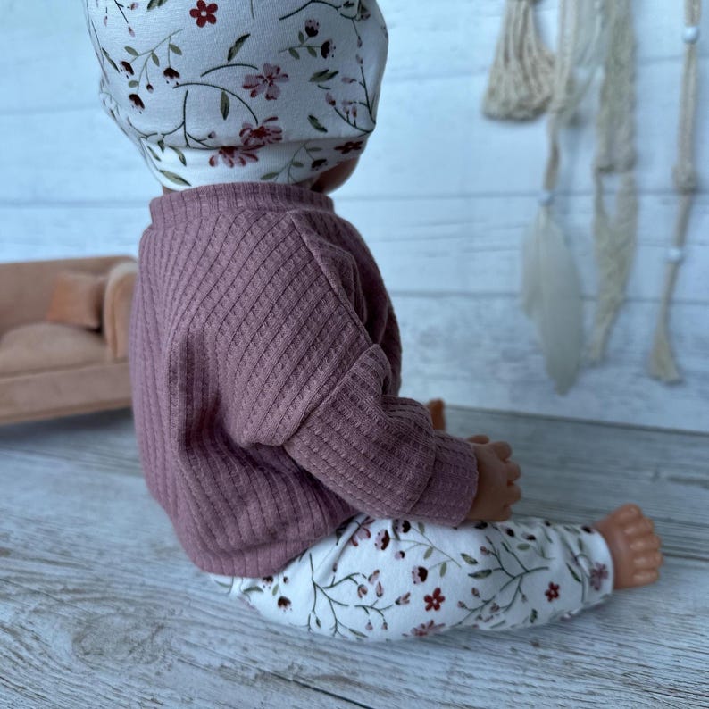 May include: A doll wearing a dusty rose ribbed long-sleeve shirt and white leggings featuring a delicate floral pattern of small red flowers and thin green vines. The doll also wears a matching white beanie with the same floral print design.