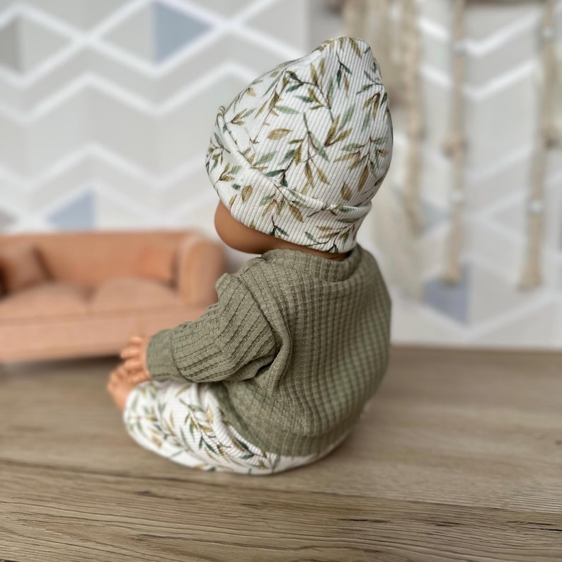 Pu&ograve; includere: Un completo per neonati composto da un berretto a coste, un maglione a maniche lunghe in maglia a nido d'ape color verde oliva e pantaloni abbinati. Il berretto e i pantaloni presentano una stampa botanica con foglie verdi e marroni su fondo bianco.