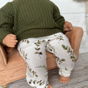 May include: A doll wearing a green ribbed sweater and white trousers with a leafy print. The doll is sitting on a light brown armchair.