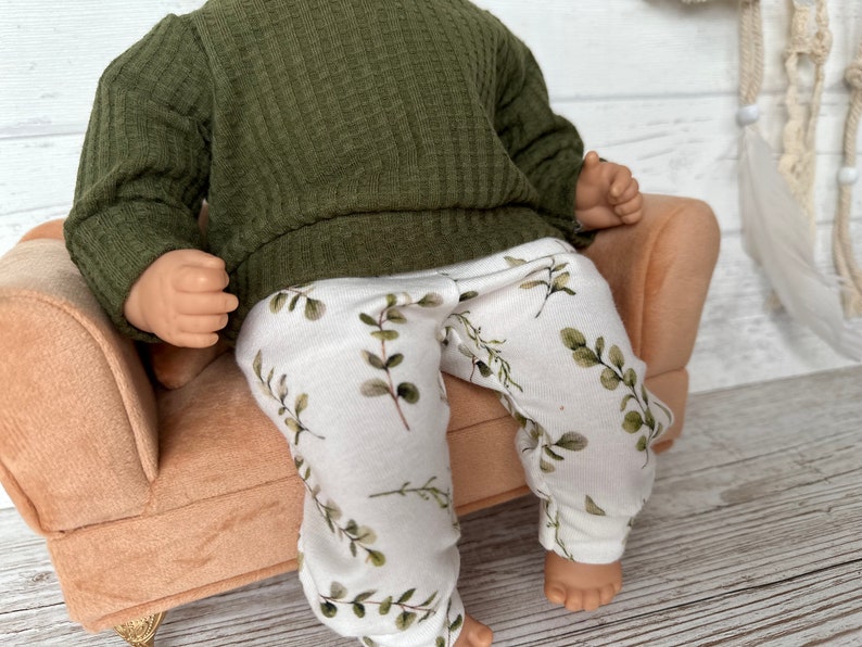 May include: A doll wearing a green ribbed sweater and white trousers with a leafy print. The doll is sitting on a light brown armchair.