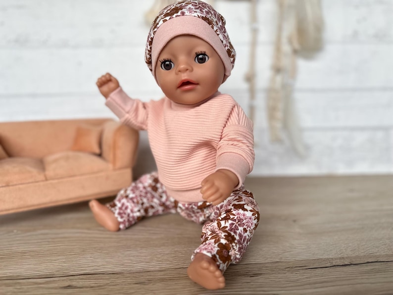 May include: A doll wearing a pink and brown floral patterned outfit with a pink beanie. The doll is sitting on a wooden surface.