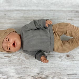 May include: A doll wearing a long-sleeved grey ribbed shirt, tan waffle-knit trousers, and a matching tan beanie. The doll has closed eyes, a small open mouth, and is positioned horizontally. The clothing features a simple, solid-coloured design.