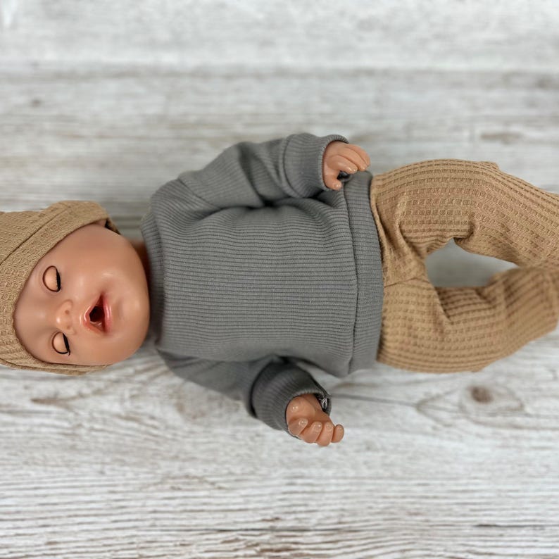 May include: A doll wearing a long-sleeved grey ribbed shirt, tan waffle-knit trousers, and a matching tan beanie. The doll has closed eyes, a small open mouth, and is positioned horizontally. The clothing features a simple, solid-coloured design.