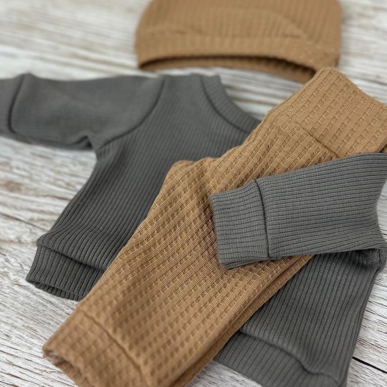 May include: A charcoal grey long-sleeve shirt with a ribbed texture, paired with tan waffle-knit trousers and a matching tan waffle-knit beanie. The garments feature cuffed sleeves and a cuffed waistband, showcasing a coordinated two-piece set.