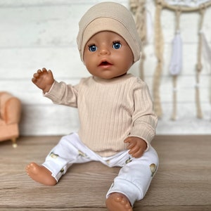 Puede incluir: Una mu&ntilde;eca beige que lleva una camisa de manga larga de punto de canal&eacute; beige y pantalones blancos con estampado floral. La mu&ntilde;eca lleva un gorro de punto beige y tiene ojos azules.