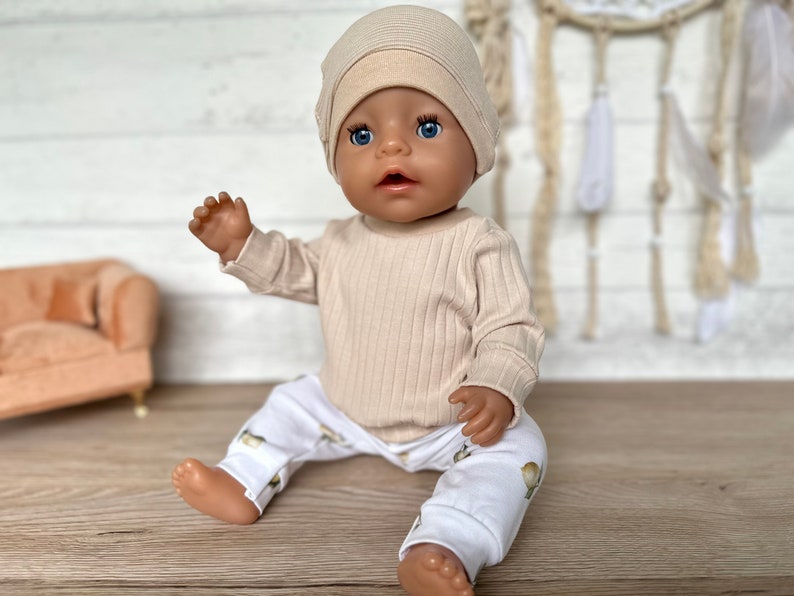 Puede incluir: Una mu&ntilde;eca beige que lleva una camisa de manga larga de punto de canal&eacute; beige y pantalones blancos con estampado floral. La mu&ntilde;eca lleva un gorro de punto beige y tiene ojos azules.