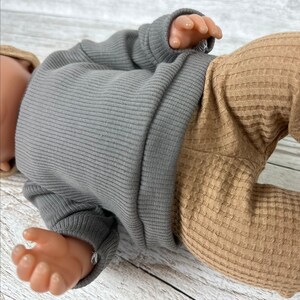 May include: A doll wearing a long-sleeved grey ribbed jumper and tan waffle-knit trousers. The doll has light skin and is positioned horizontally, showing the texture of the fabric on the jumper and the trousers clearly in the frame.