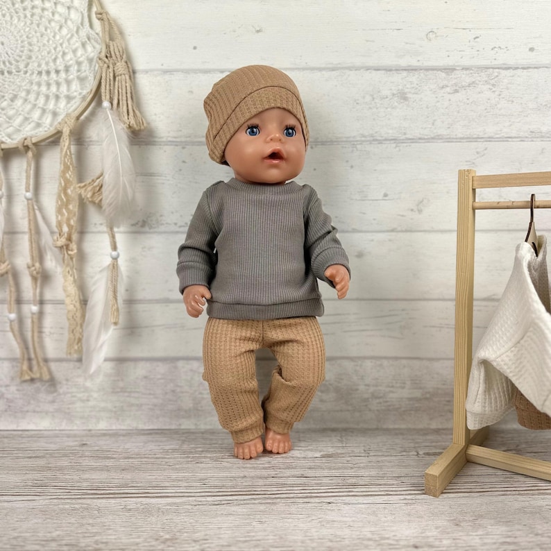 May include: A doll with blue eyes wearing a tan beanie, a long-sleeved grey ribbed shirt, and tan waffle-knit trousers. The doll stands upright, featuring a neutral expression, positioned against a light-coloured wooden background with a dreamcatcher.