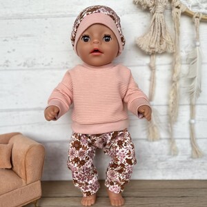 May include: A doll wearing a pink sweater and floral print trousers with a floral print hat. The doll has brown eyes and dark brown hair.