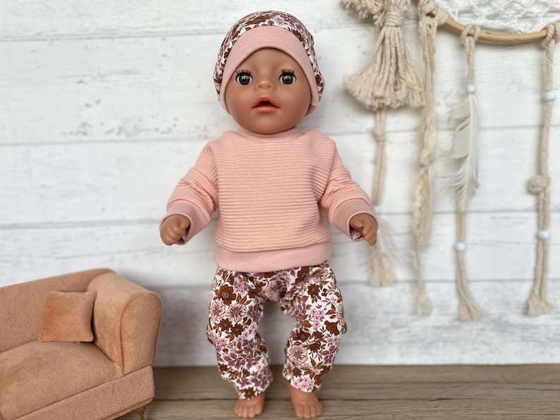 May include: A doll wearing a pink sweater and floral print trousers with a floral print hat. The doll has brown eyes and dark brown hair.