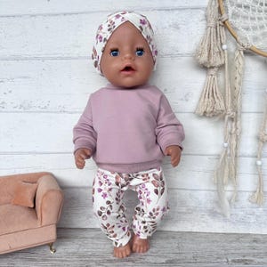 May include: A doll with blue eyes wearing a mauve long-sleeve sweatshirt and white leggings featuring a pattern of brown leaves and small purple flowers. The doll also wears a matching white head wrap with the same leaf and flower print design.