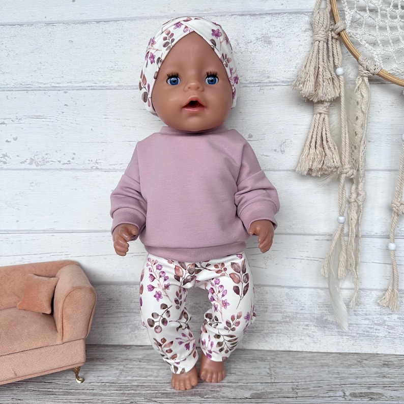 May include: A doll with blue eyes wearing a mauve long-sleeve sweatshirt and white leggings featuring a pattern of brown leaves and small purple flowers. The doll also wears a matching white head wrap with the same leaf and flower print design.
