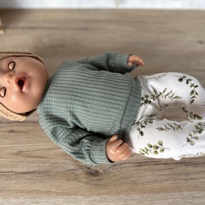 May include: A doll wearing a green waffle knit long-sleeve shirt and white pants with a green leaf pattern. The doll is wearing a tan knitted hat.