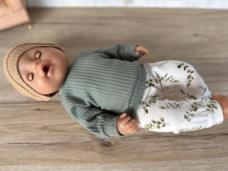 May include: A doll wearing a green waffle knit long-sleeve shirt and white pants with a green leaf pattern. The doll is wearing a tan knitted hat.