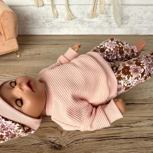 May include: A pink ribbed sweater and floral print trousers for a doll. The doll is wearing a floral print hat.