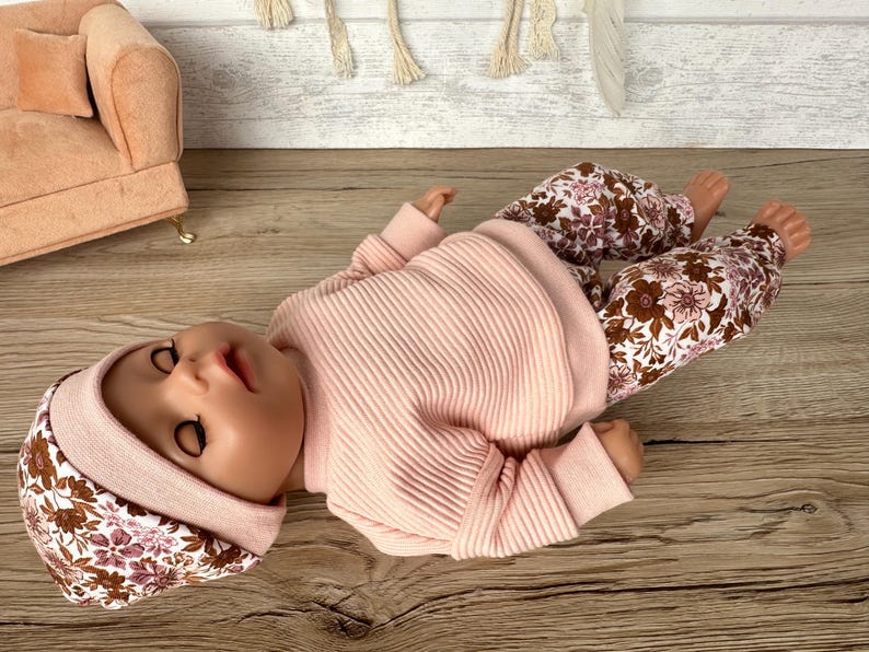 May include: A pink ribbed sweater and floral print trousers for a doll. The doll is wearing a floral print hat.