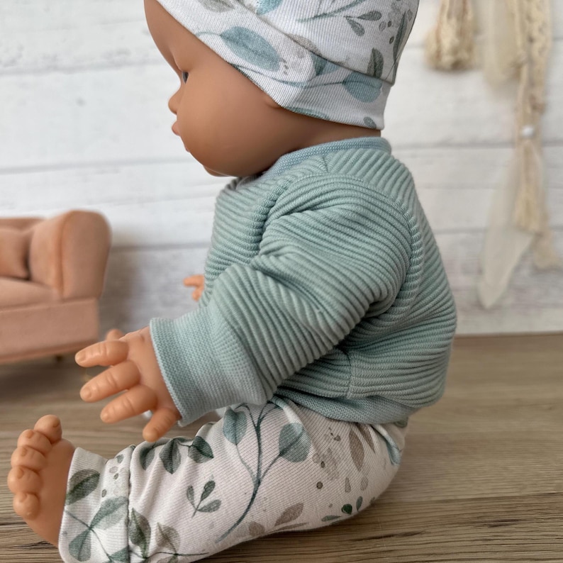 May include: A doll wearing a sage green ribbed long-sleeve shirt, white leggings with a green and brown botanical leaf print, and a matching white beanie with the same leaf pattern. The doll has a smooth, light brown complexion and is seated.