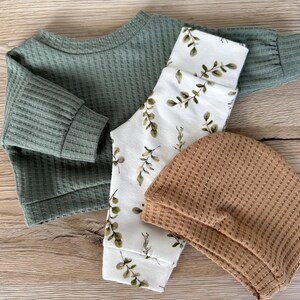 May include: A set of baby clothes in green, white, and brown. The set includes a long-sleeved shirt, pants with a leafy print, and a beanie.