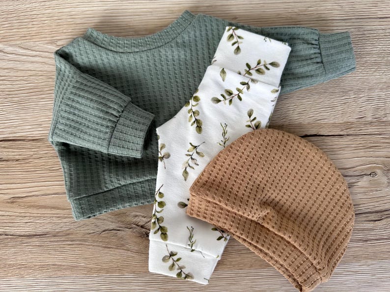 May include: A set of baby clothes in green, white, and brown. The set includes a long-sleeved shirt, pants with a leafy print, and a beanie.