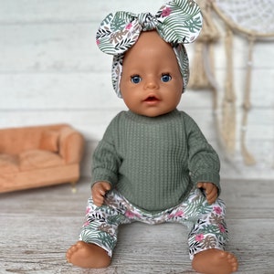 May include: A doll wearing a green waffle knit long-sleeve shirt and floral print pants. The doll is wearing a floral print headband with a bow.