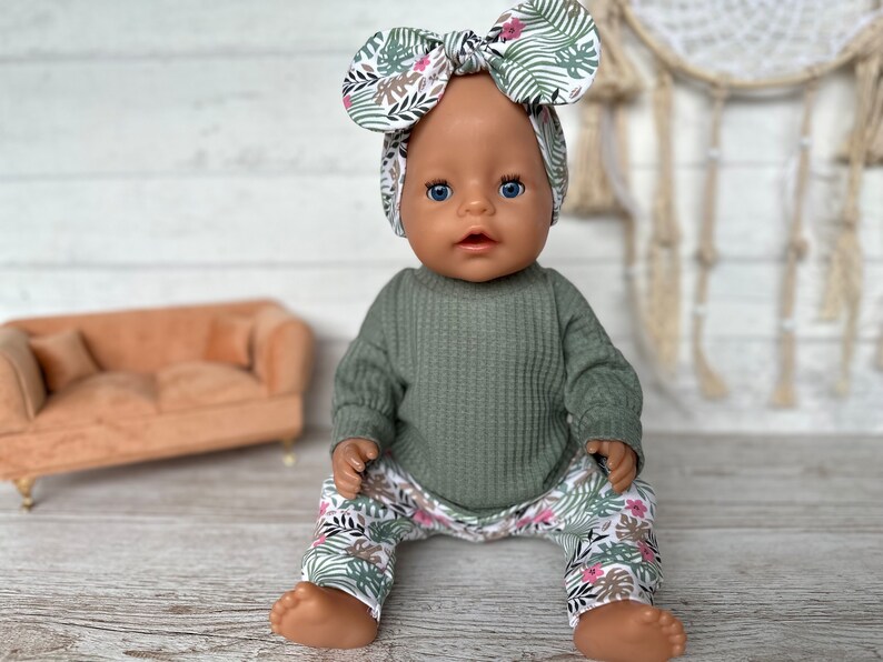 May include: A doll wearing a green waffle knit long-sleeve shirt and floral print pants. The doll is wearing a floral print headband with a bow.