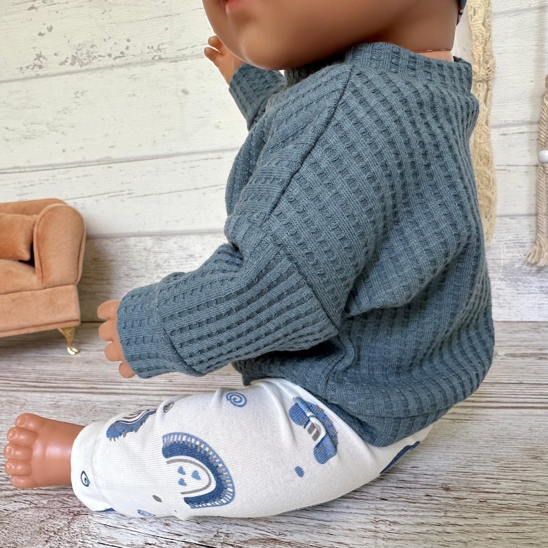 May include: A doll wearing a long-sleeved, slate blue waffle-knit sweater and white leggings featuring a repeating pattern of blue rainbows and small hearts. The doll is positioned in a seated posture, showcasing the textured fabric of the top.