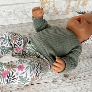 May include: A baby doll wearing a green long-sleeved shirt and white pants with a tropical leaf and flower print. The doll is wearing a matching headband with the same print.