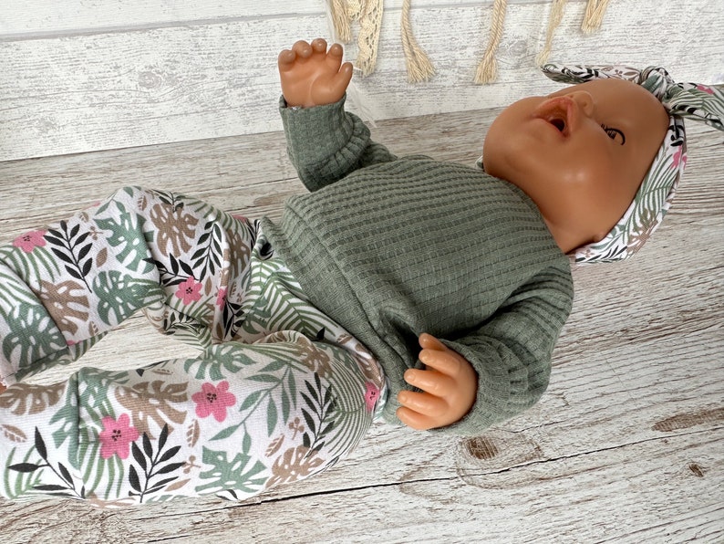 May include: A baby doll wearing a green long-sleeved shirt and white pants with a tropical leaf and flower print. The doll is wearing a matching headband with the same print.