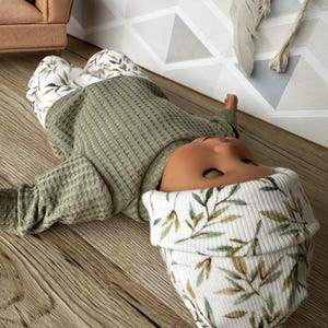 Pu&ograve; includere: Una bambola indossa una maglia a maniche lunghe in tessuto a nido d'ape verde oliva, pantaloni bianchi con motivo a foglie verdi e marroni e un berretto bianco abbinato con motivo a foglie. La bambola &egrave; sdraiata sulla schiena con la testa girata.