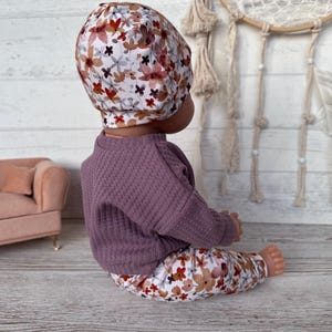 May include: A baby doll wearing a floral print hat, trousers, and a purple waffle-knit sweater. The floral pattern includes red, brown, and white flowers. The doll is sitting on a light wood surface.