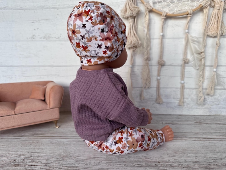 May include: A baby doll wearing a floral print hat, trousers, and a purple waffle-knit sweater. The floral pattern includes red, brown, and white flowers. The doll is sitting on a light wood surface.