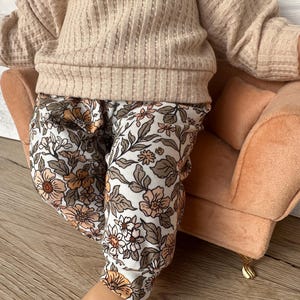 May include: A baby wearing a beige sweater and floral print pants sits on a peach-colored armchair. The armchair has a gold-colored base.