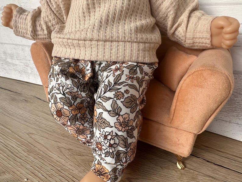 May include: A baby wearing a beige sweater and floral print pants sits on a peach-colored armchair. The armchair has a gold-colored base.