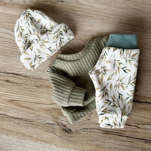 Pu&ograve; includere: Un set di abbigliamento in maglia a nido d'ape composto da un berretto, una maglia a maniche lunghe e un pantalone. Il berretto e il pantalone presentano uno sfondo bianco con un motivo a foglie verdi e marroni. La maglia &egrave; verde salvia tinta unita.