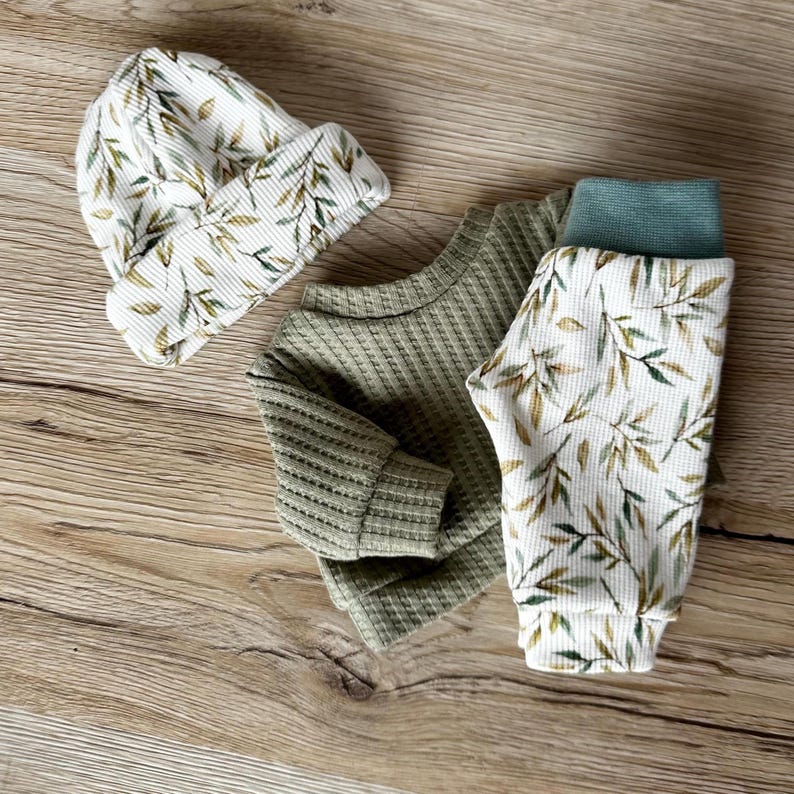 Pu&ograve; includere: Un set di abbigliamento in maglia a nido d'ape composto da un berretto, una maglia a maniche lunghe e un pantalone. Il berretto e il pantalone presentano uno sfondo bianco con un motivo a foglie verdi e marroni. La maglia &egrave; verde salvia tinta unita.