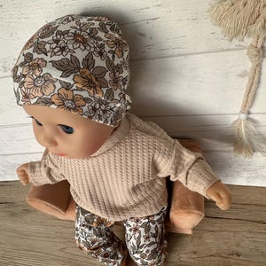May include: A doll wearing a beige waffle knit sweater and floral print pants. The doll is wearing a white floral print beanie and is sitting on a brown cushion.