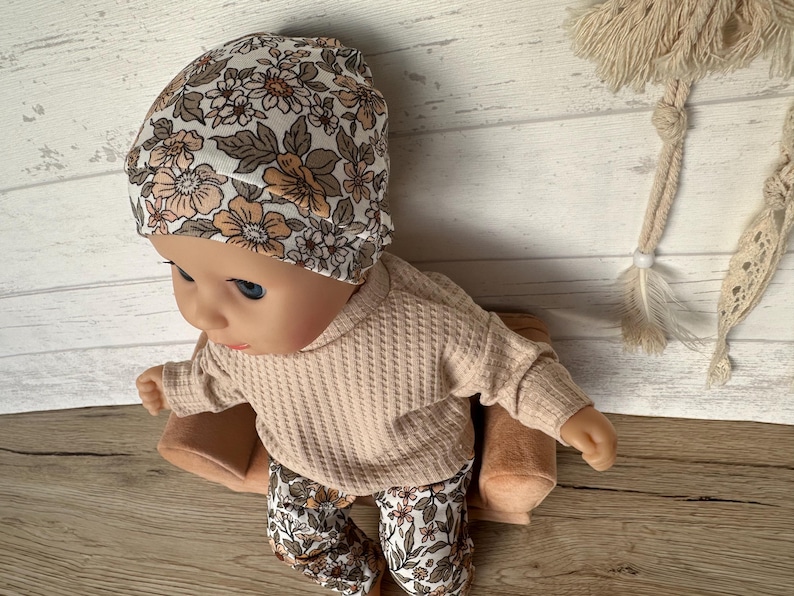 May include: A doll wearing a beige waffle knit sweater and floral print pants. The doll is wearing a white floral print beanie and is sitting on a brown cushion.