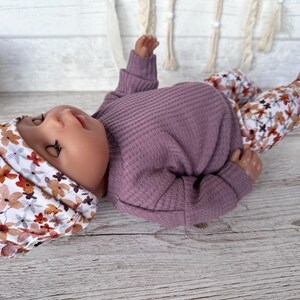 May include: A doll dressed in a mauve waffle-knit top, floral print trousers, and a matching floral print beanie. The trousers and beanie have a white background with brown, orange, and mauve flowers.