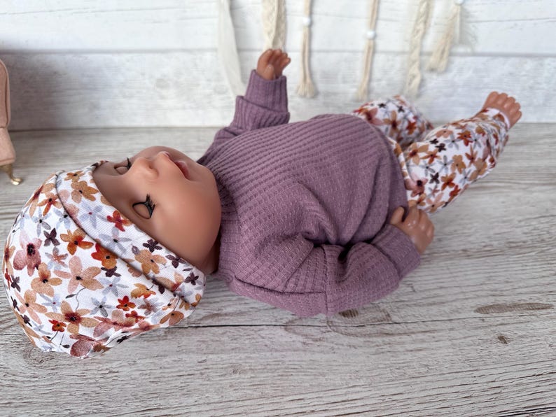 May include: A doll dressed in a mauve waffle-knit top, floral print trousers, and a matching floral print beanie. The trousers and beanie have a white background with brown, orange, and mauve flowers.